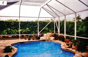 glass enclosed swimming pool with hardscape waterfall and plants on the pool deck