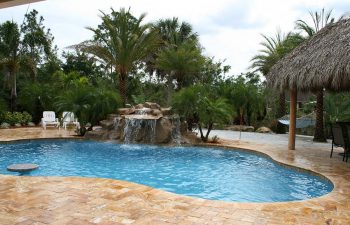 a backyard swimming pool with hardscape waterfall and tiki hut on a Travertine deck