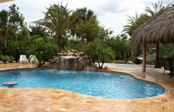 a backyard swimming pool with waterfall and tiki hut on a Travertine deck