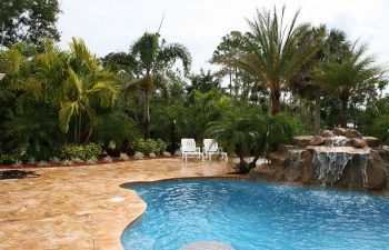 a backyard swimming pool with waterfall and tiki hut on a Travertine deck