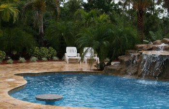 a backyard swimming pool with waterfall and tiki hut on a Travertine deck