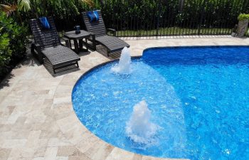 two sunbeds and a table by a backyard swimming pool with built-in fountains