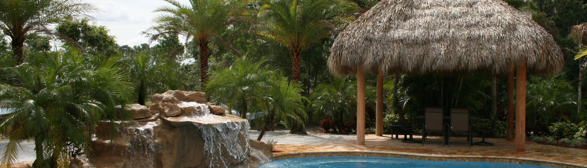 a hardscape waterfall and a tiki hut on deck of a backyard swimming pool